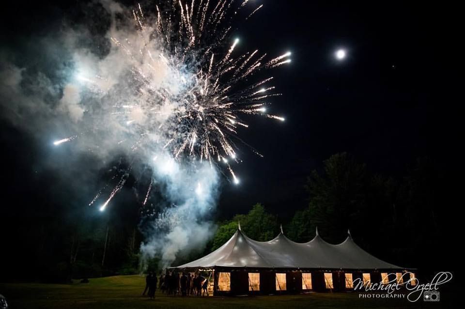 Fireworks burst above a lit tent at night; people stand near tent entrance.