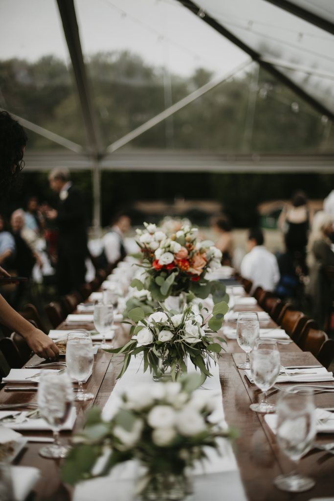 Long wooden tables set for a wedding reception under a clear tent; floral centerpieces, guests in background.