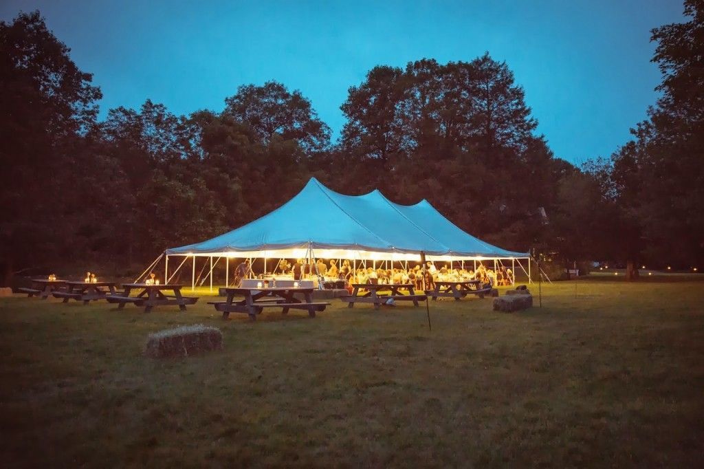 White tent lit up at dusk, picnic tables on grass, surrounded by trees.