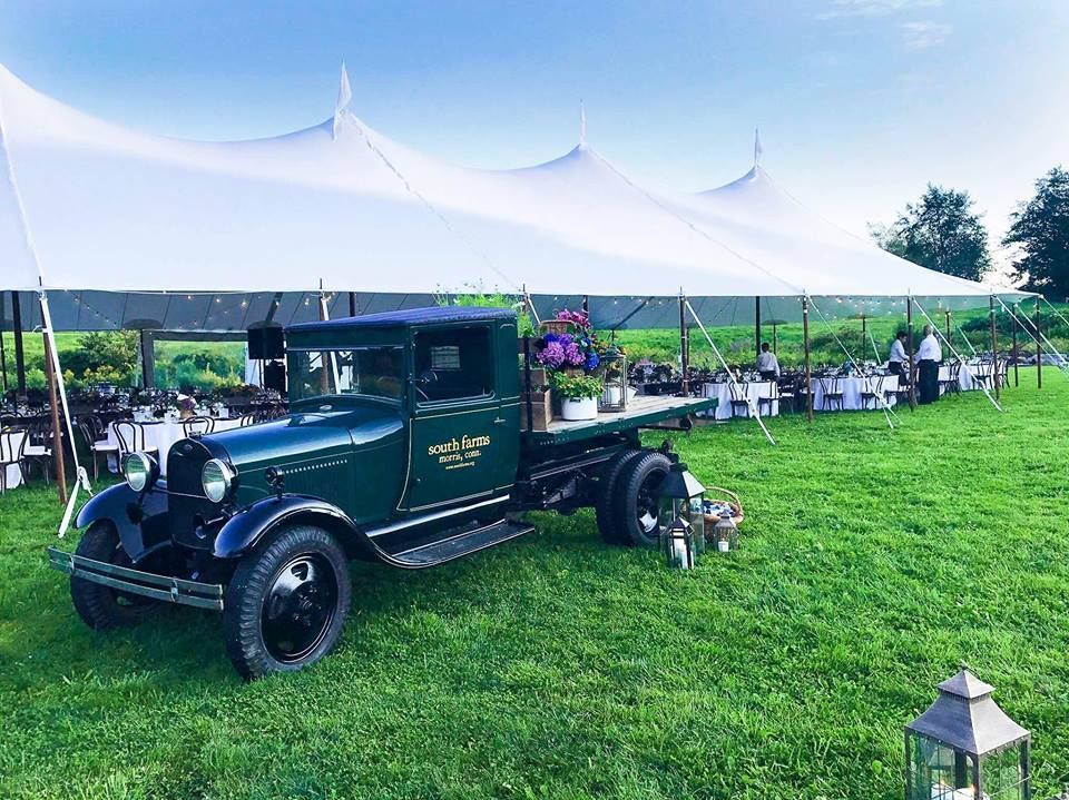Green vintage truck with floral display, set up as a bar next to a large white tent at an outdoor event.