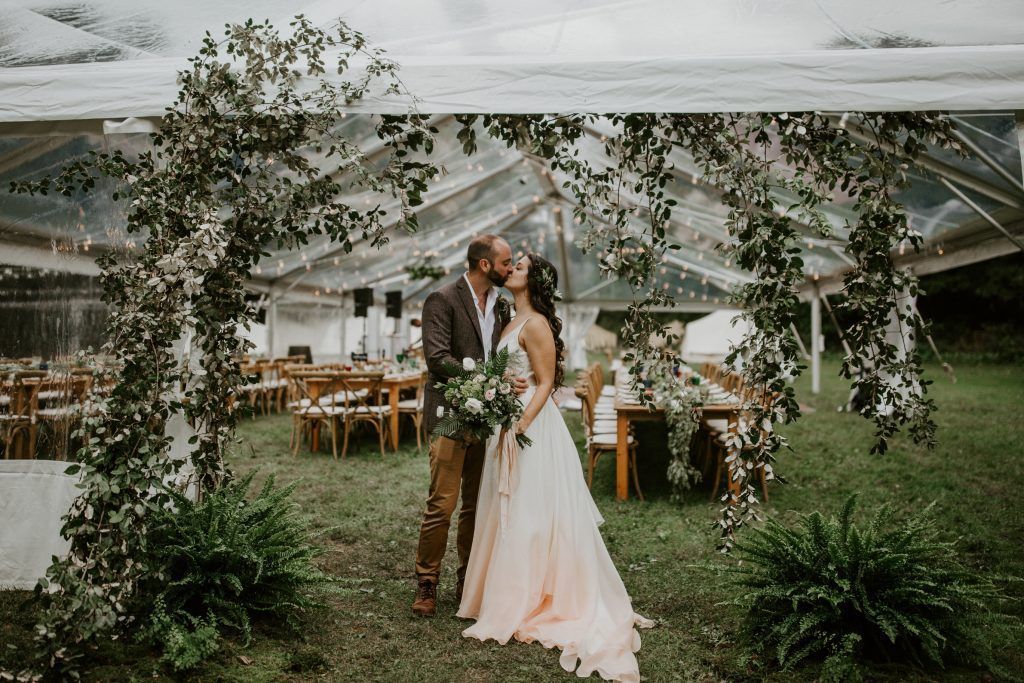 Couple kissing under floral arch at outdoor wedding reception.