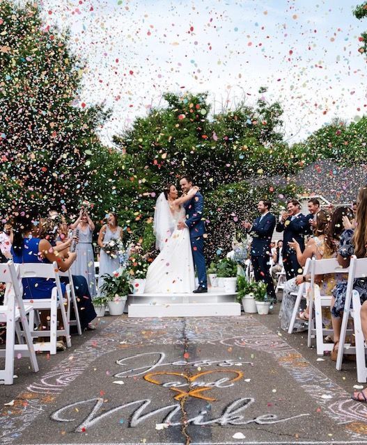 Couple kissing under confetti at outdoor wedding ceremony. Guests watch; flower decorations.