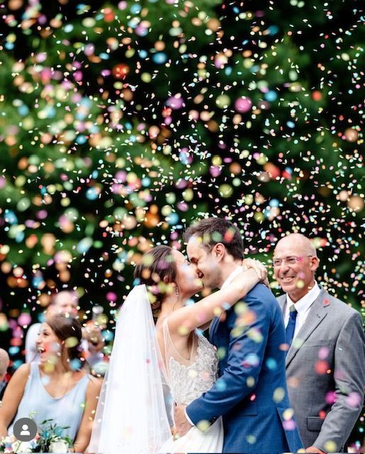 Couple kisses amidst confetti at outdoor wedding ceremony.