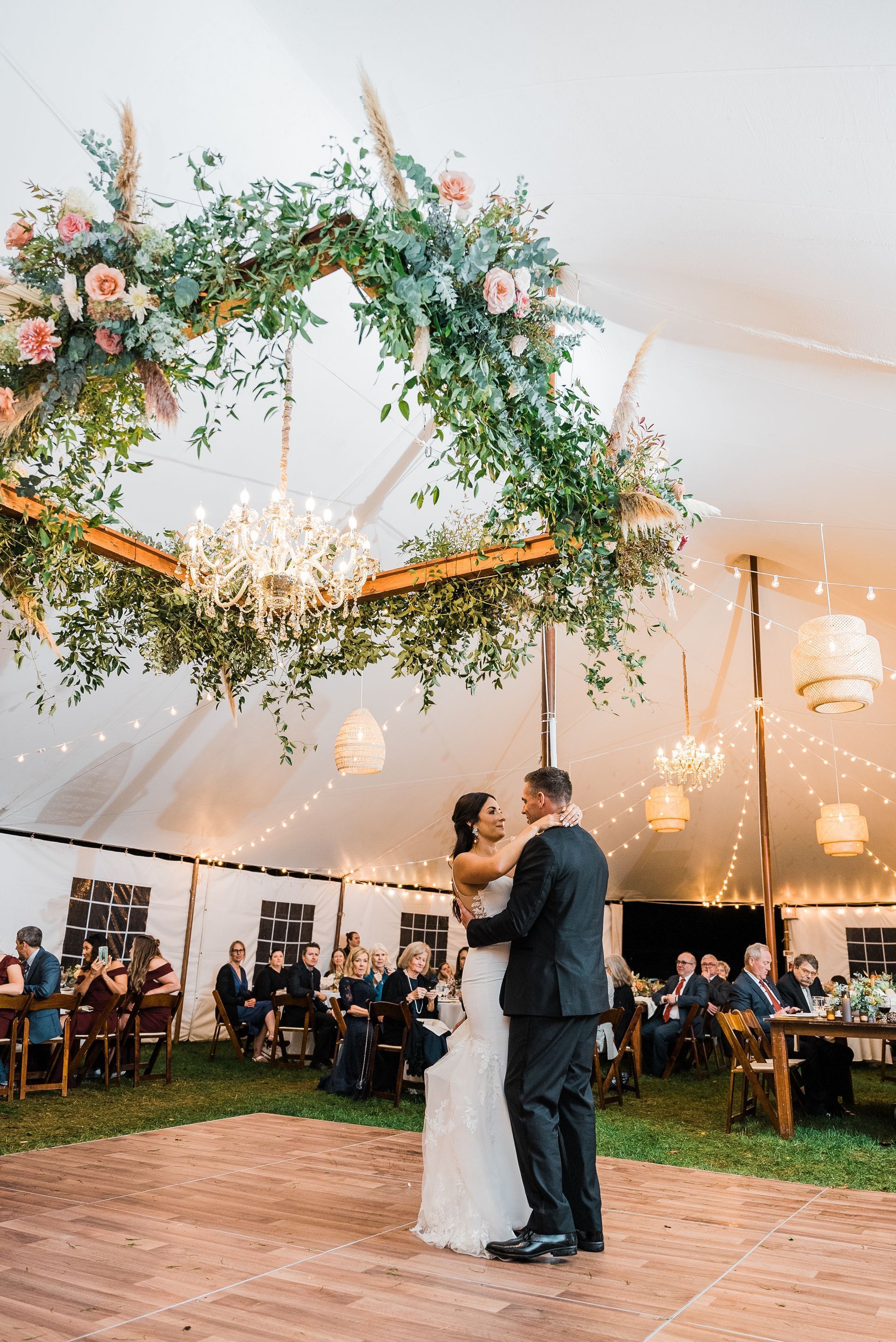 Couple dancing at a wedding reception under a floral arch and tent, guests seated at tables.