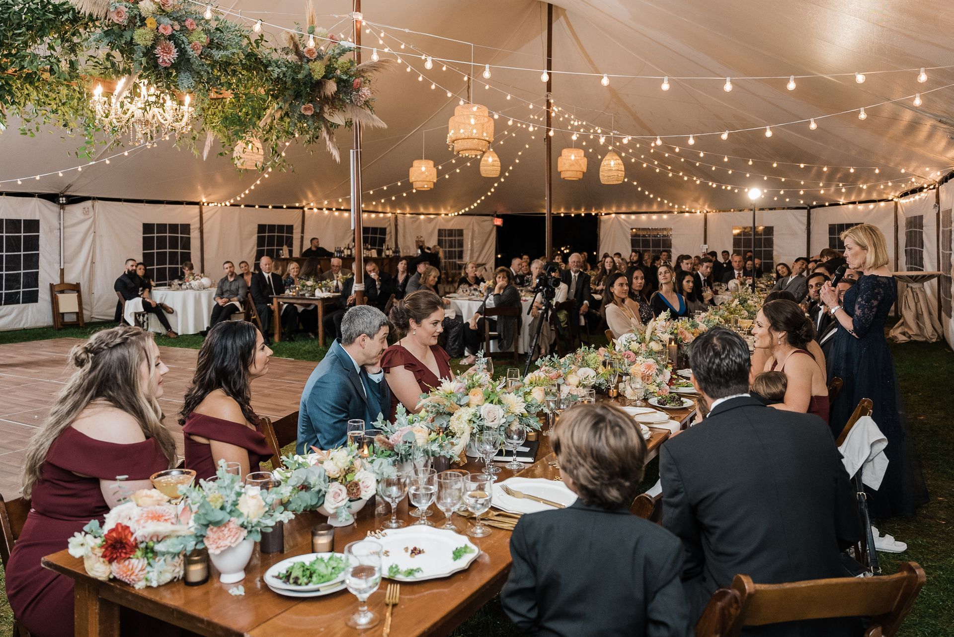 Wedding reception in a tent, tables set with flowers and guests seated, overhead string lights.