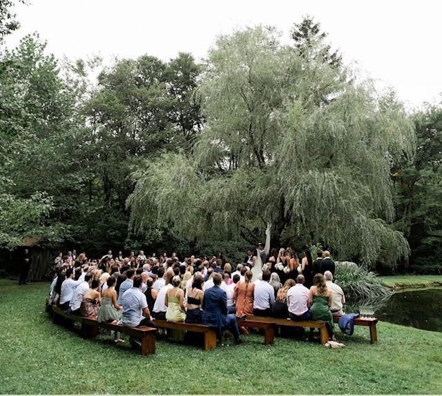 Wedding ceremony outdoors under a large willow tree. Guests seated on benches face a couple and officiant.