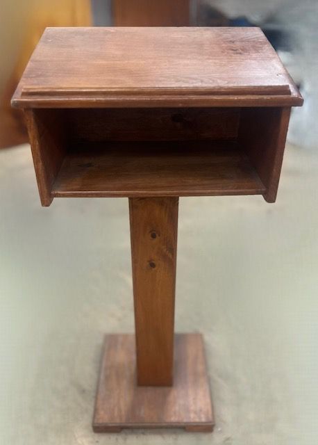 Wooden lectern with a rectangular top and square base, set against a blurred background.