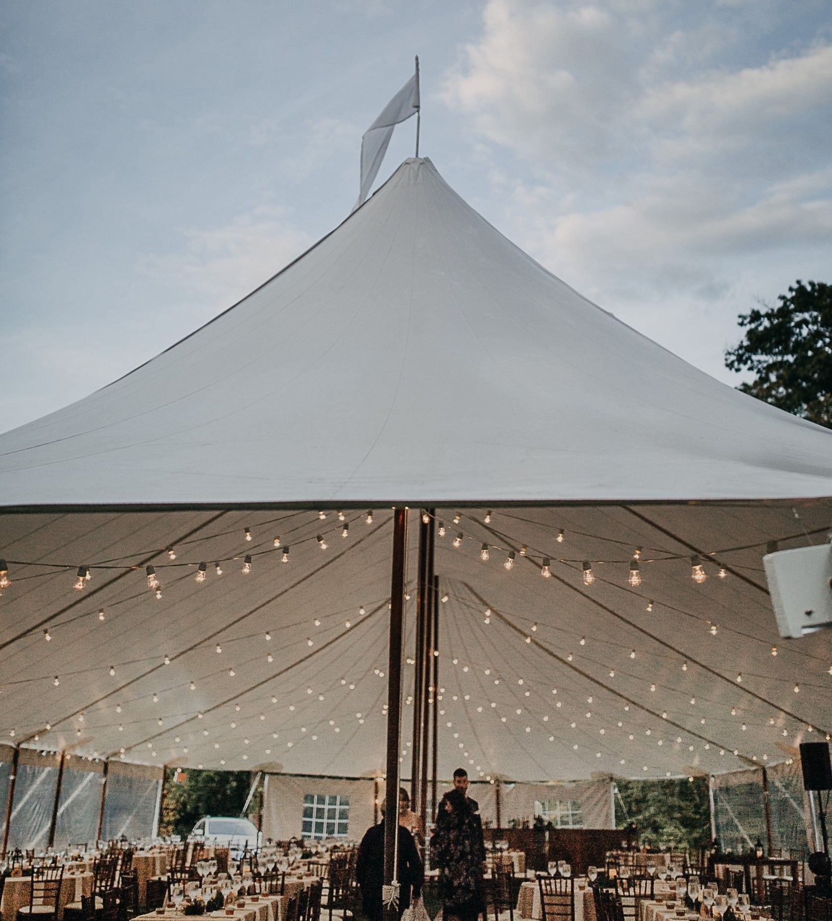 White tent with string lights over a reception. People stand at tables inside. Flag at the top.