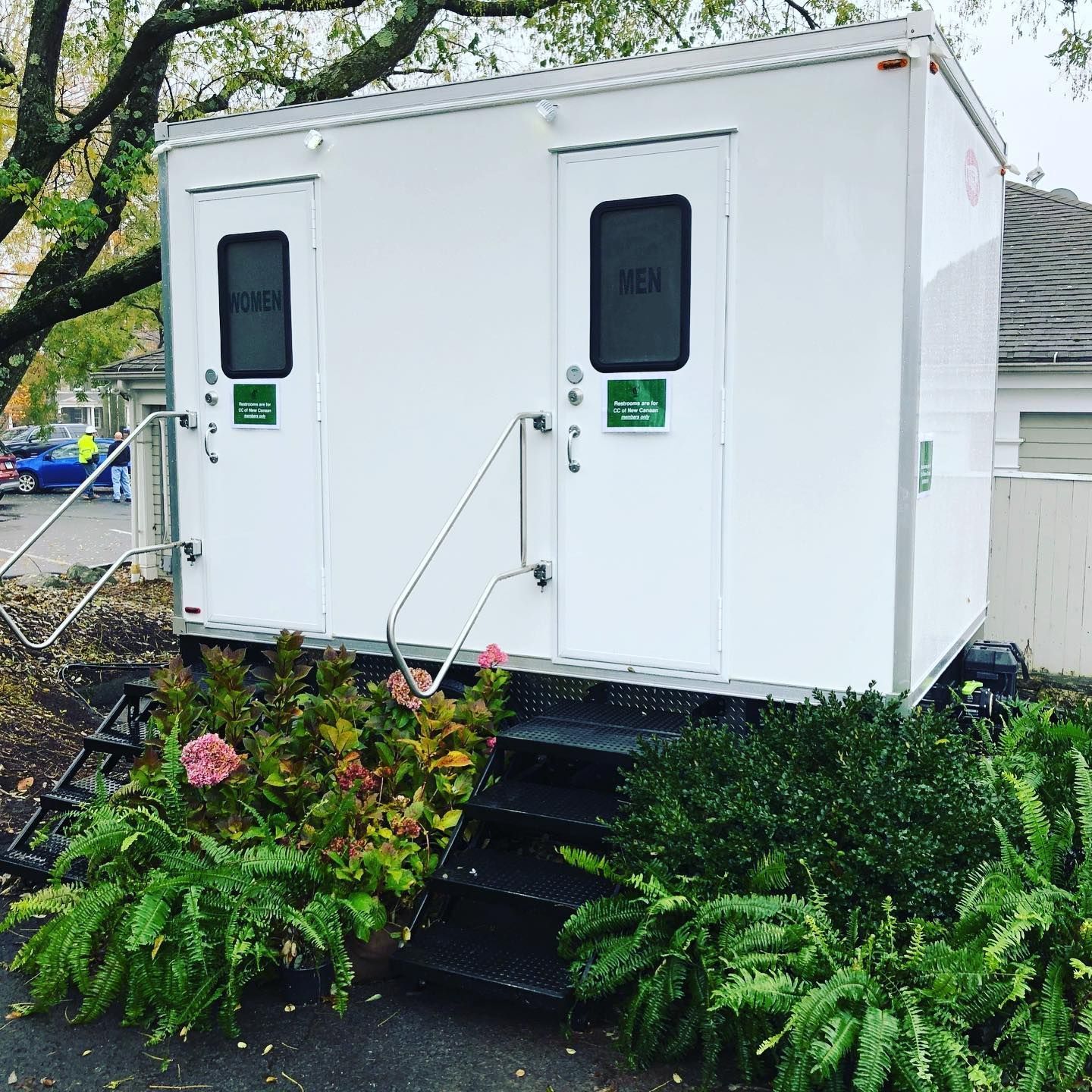White portable restroom trailer with men's and women's entrances, set on a landscaped area with stairs.