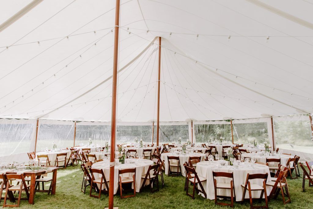 Tent set up for a wedding reception, with round tables, chairs, and string lights on a grassy lawn.