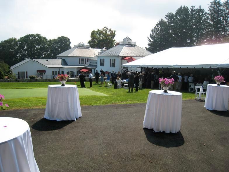 Outdoor event: White-clothed tables with flower centerpieces on a black surface; white tent; people gather on grass in front of white building.