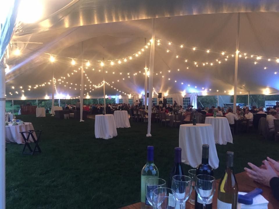 Large tent party with string lights and white table covers; people seated at tables, wine bottles in foreground.
