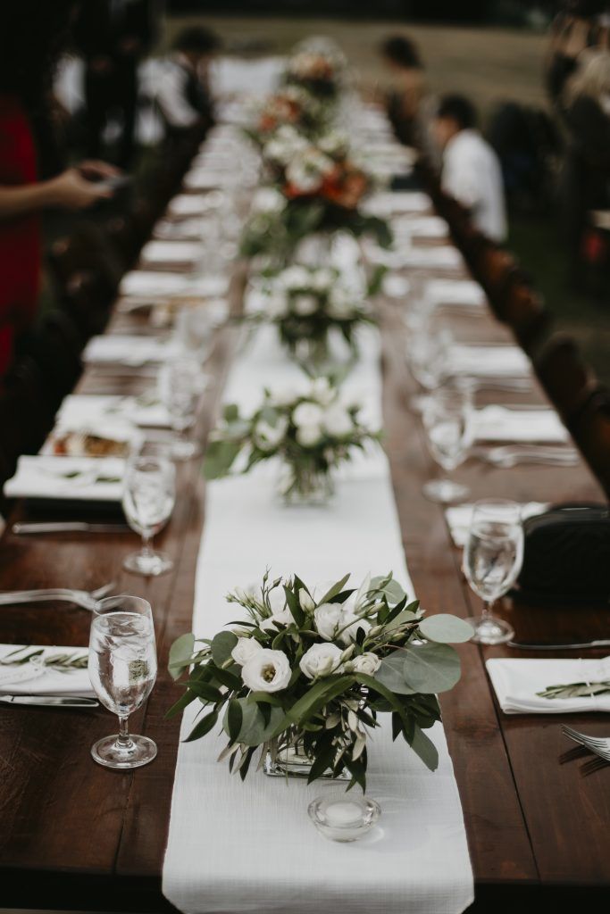 Long wooden table set for a formal outdoor event, with white floral centerpieces and glassware.