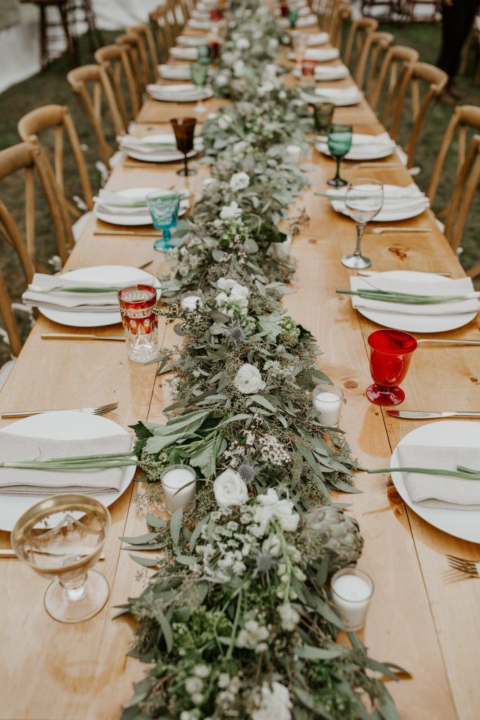 Long wooden dining table set for a celebration, decorated with greenery and colorful glassware.