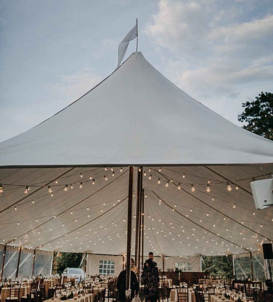White tent, lit by string lights, set for a celebration. People gather inside.
