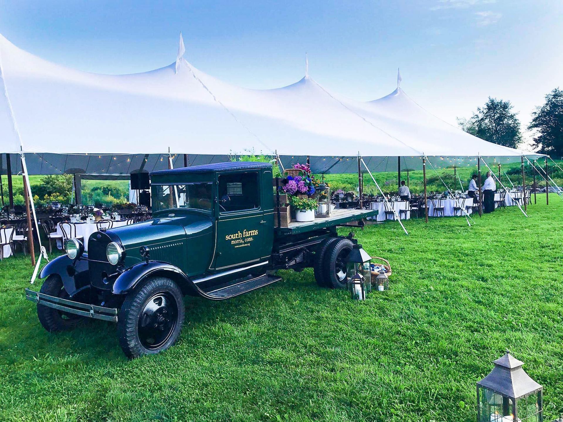 Green vintage truck with flowers, in a field, serving as a bar, next to a white tent at an outdoor event.