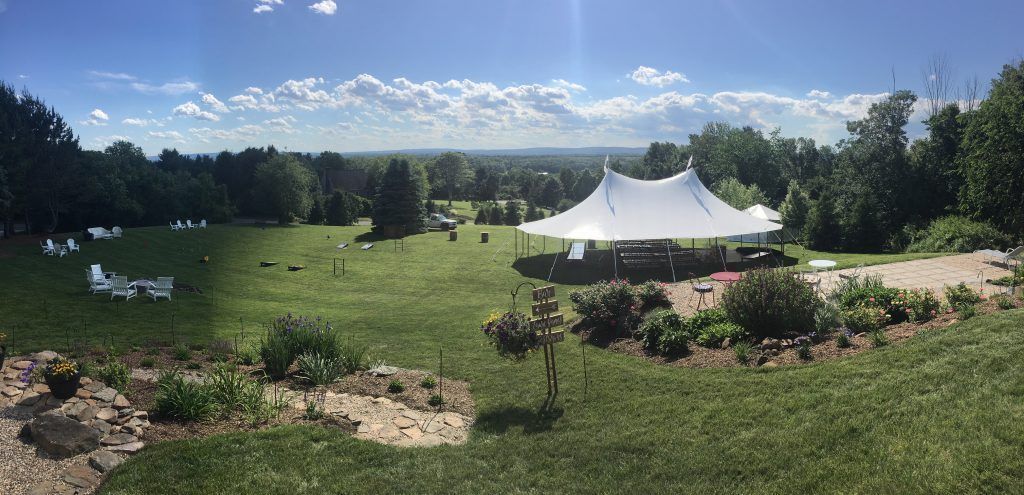 Wide shot of a grassy field with a large white tent. Trees and buildings are visible in the background under a blue sky.