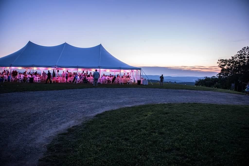 Tent illuminated at dusk with people inside, set on a grassy hill with a gravel path, overlooking a distant horizon.