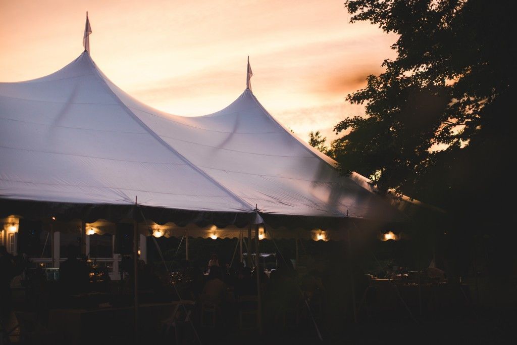 White tent at dusk with people inside, lit by soft lights; sunset in background.