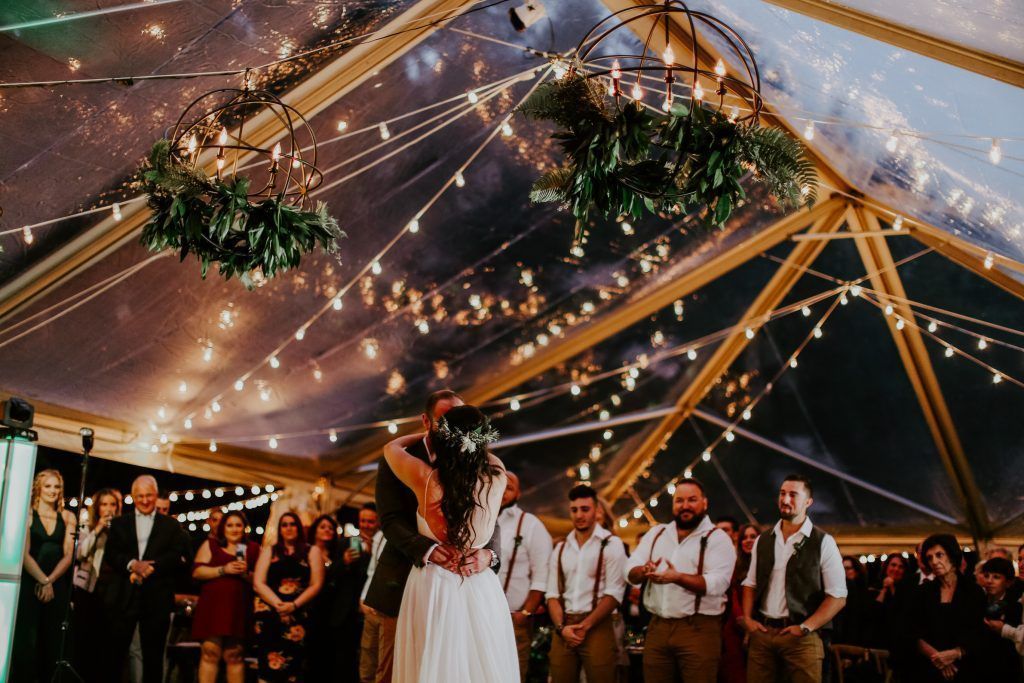 Wedding couple dancing under string lights in clear tent, guests watching.