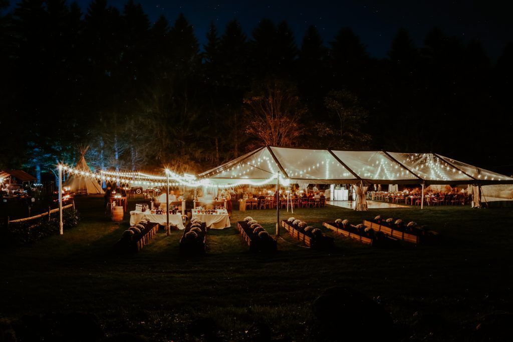 Nighttime outdoor wedding reception under a lit, transparent tent. Tables and barrels are set up, with string lights overhead.