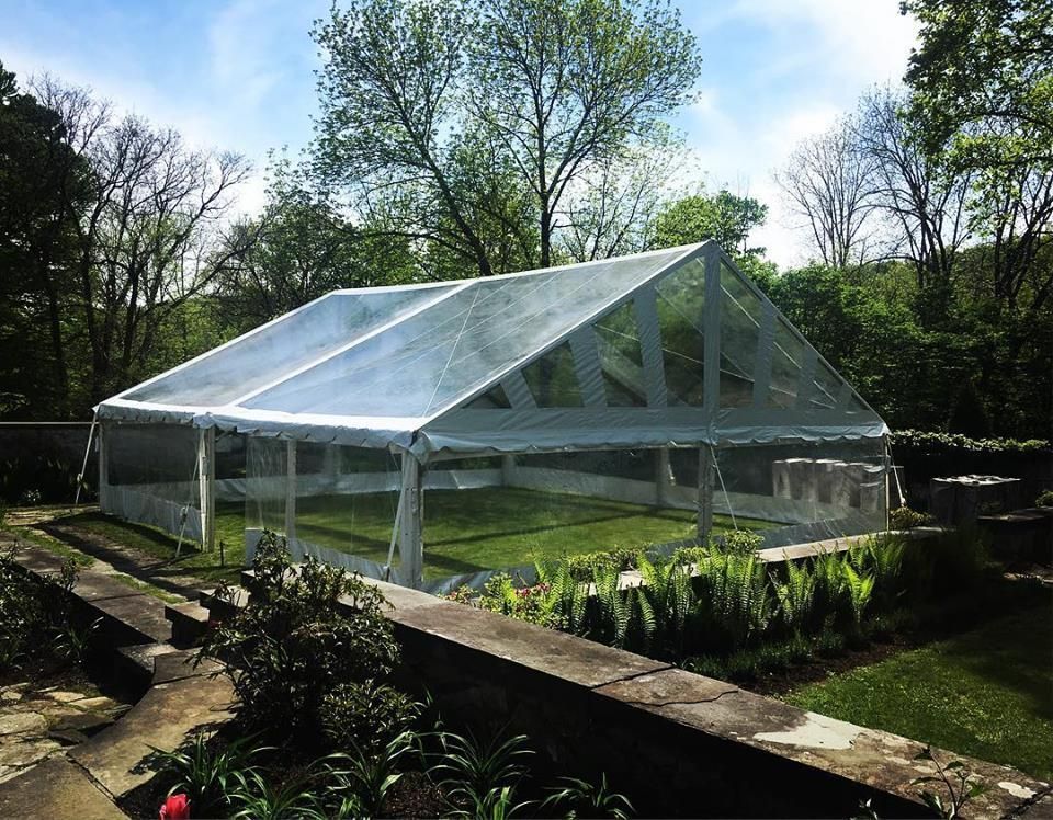 Clear tent set up on green lawn, surrounded by trees and shrubs.