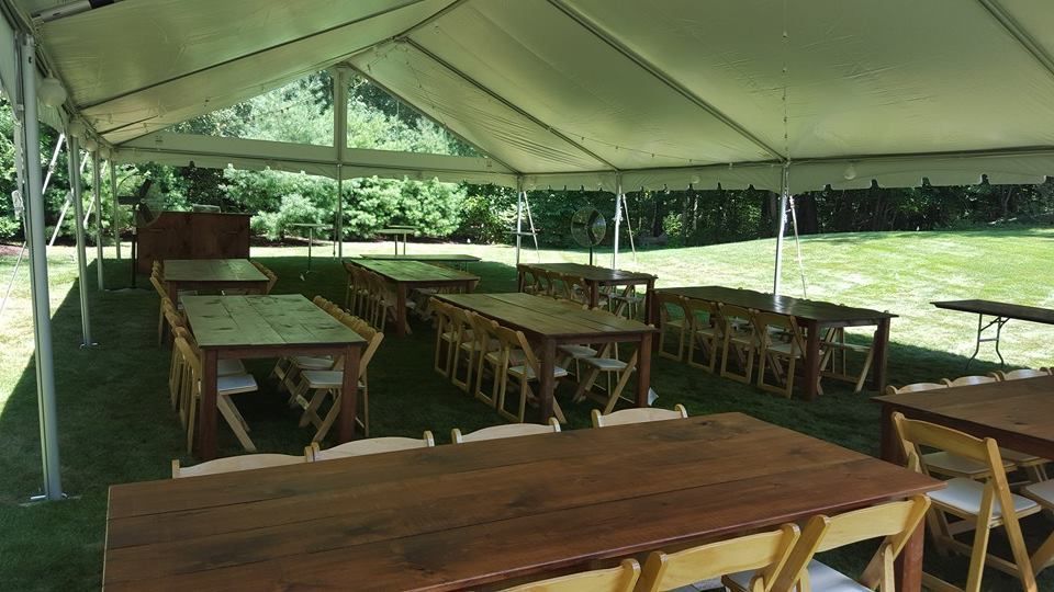 Tented outdoor event space with long wooden tables and folding chairs set on a grassy lawn.