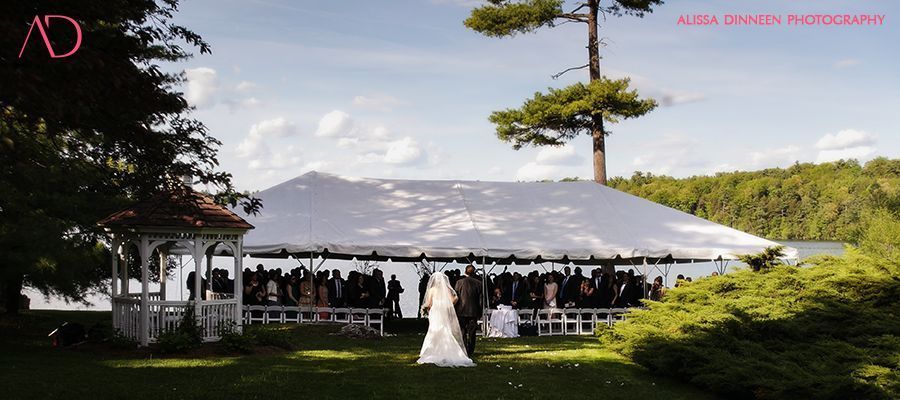 Wedding ceremony with couple walking towards tent, guests seated outside, greenery and water in background.