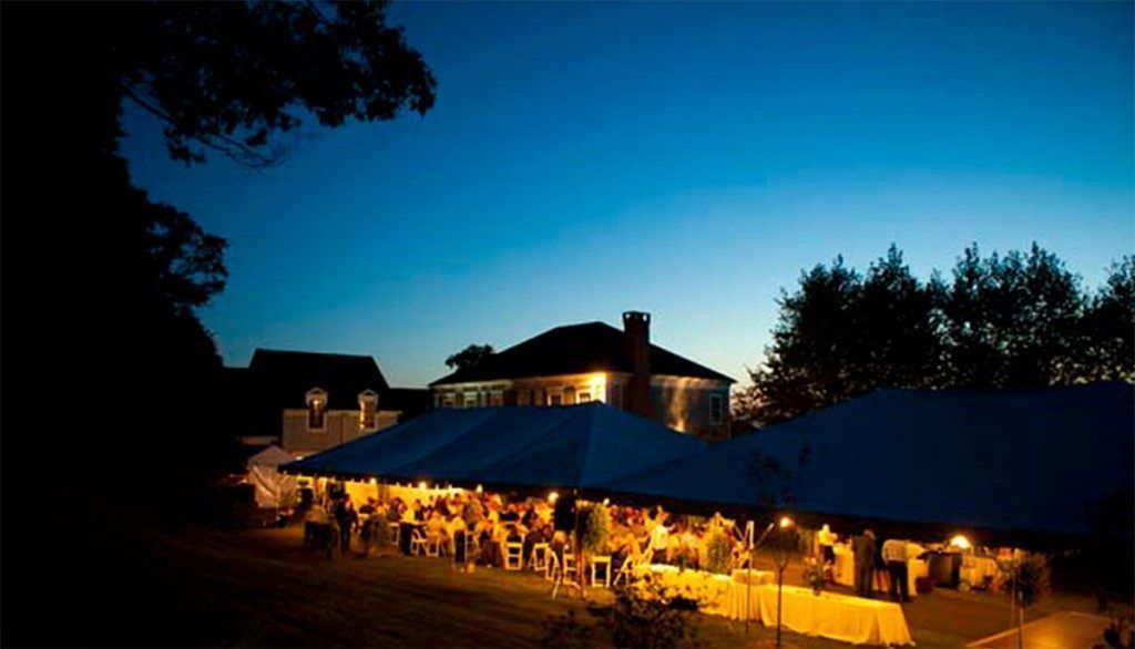 Evening outdoor event under a long tent. Tables and guests are lit, with a house and trees in the background.