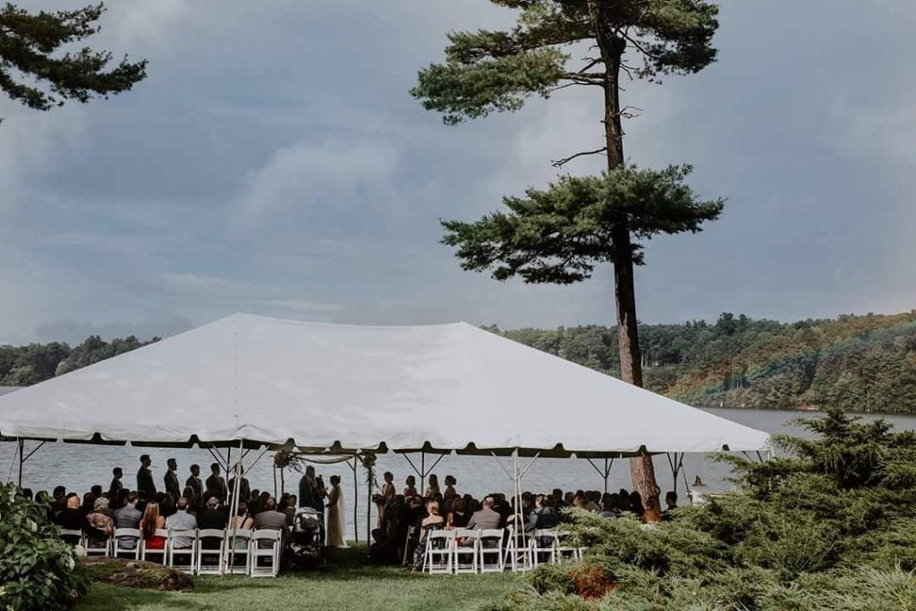 Wedding ceremony under white tent near lake; guests seated in white chairs, overcast sky.