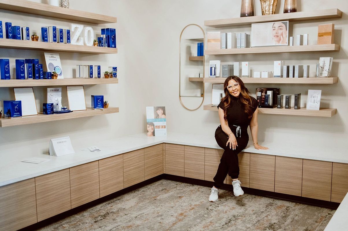 Woman sitting in a skincare product display room, smiling. Shelves with skincare products. Beige and white tones.