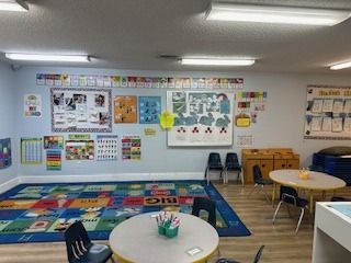 A classroom with tables, chairs, colorful rug, educational posters, and children's toys.
