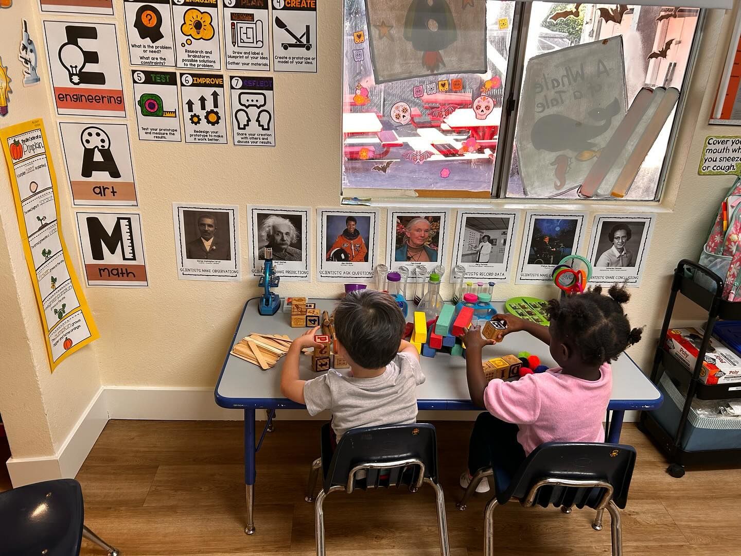 Two children playing at a small table in a classroom with educational posters and a window.