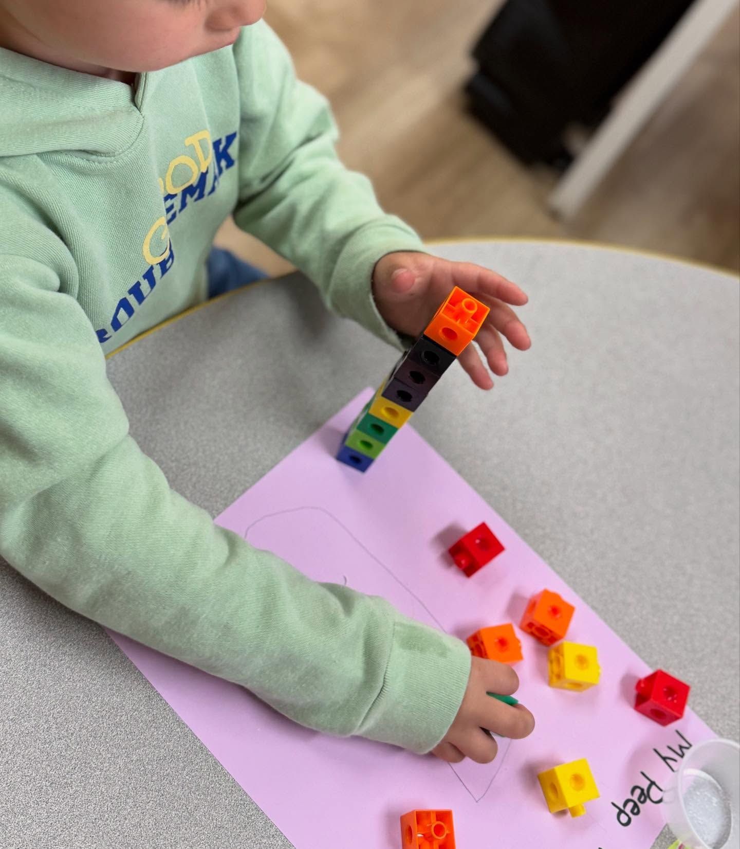Child stacking colorful blocks on a paper cutout, surrounded by scattered blocks.