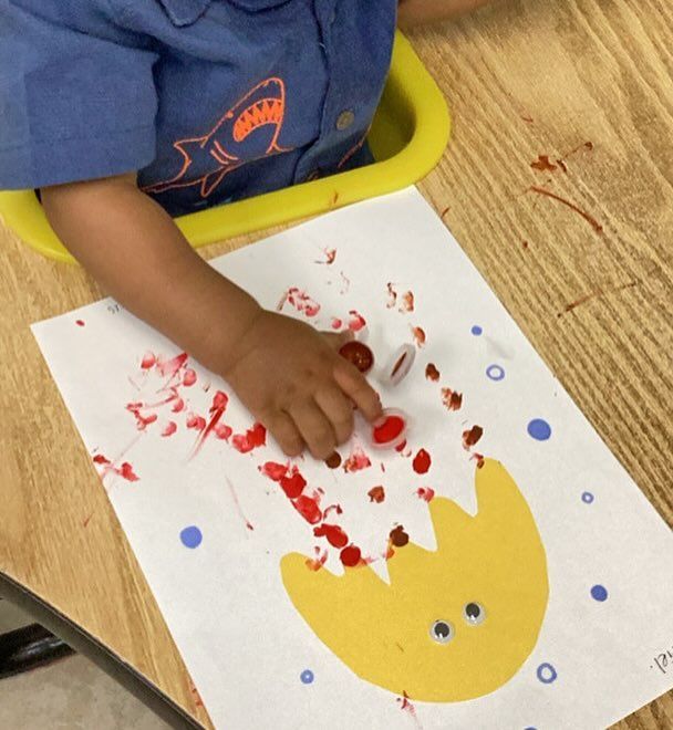 Child painting with red paint on a paper egg shape at a table.