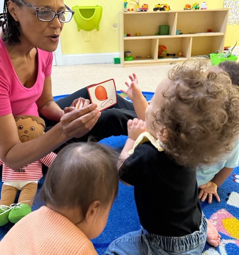Teacher reading a card to toddlers in a colorful classroom.