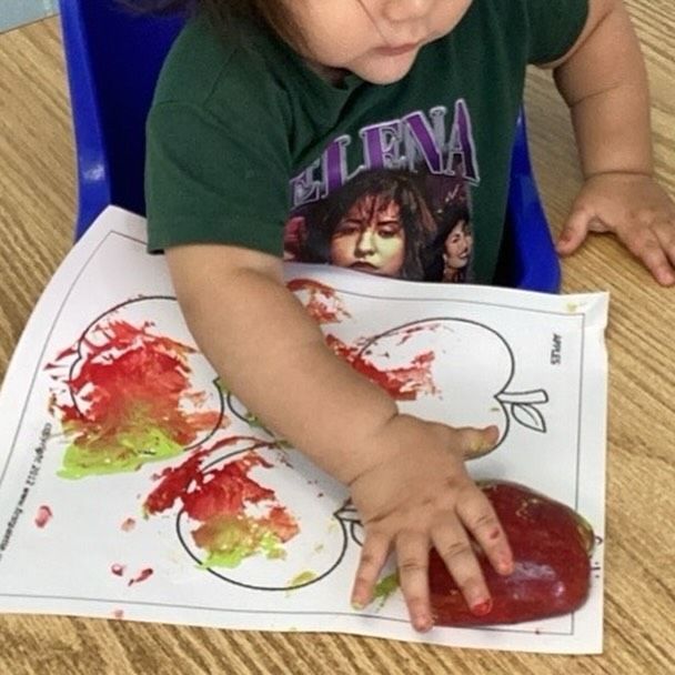 Child using an apple to paint on a worksheet with apple outlines. Red and green paint are visible.