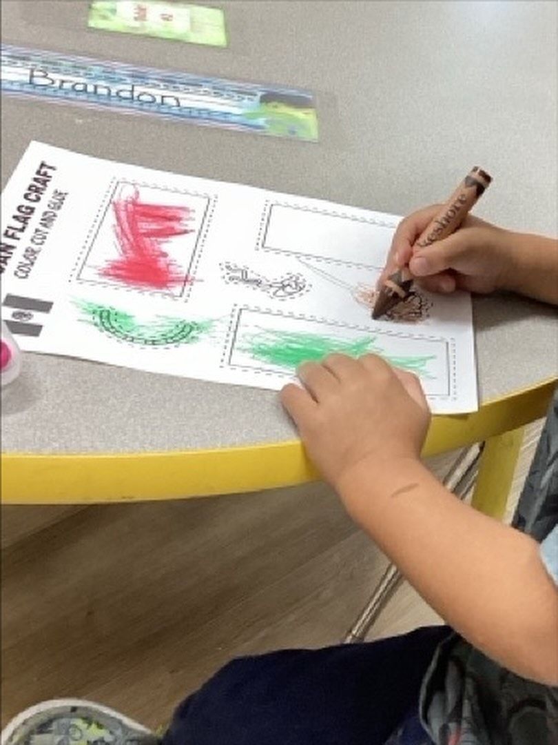 Child coloring a flag craft with brown crayon at a table.