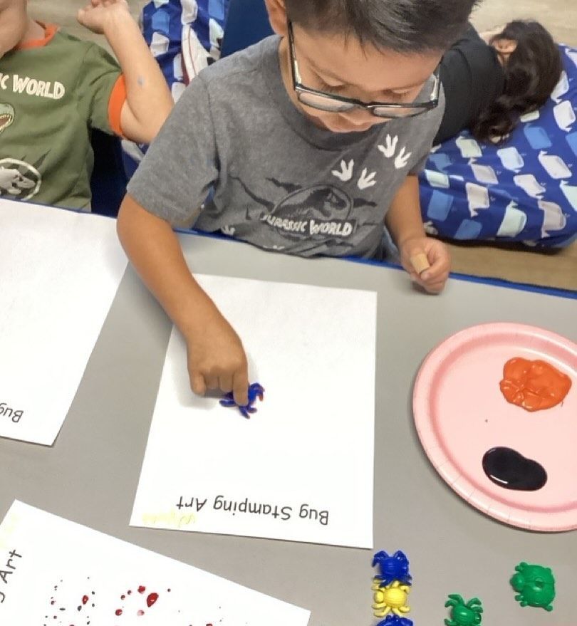 Boy with glasses stamping paint bugs on paper at a table.