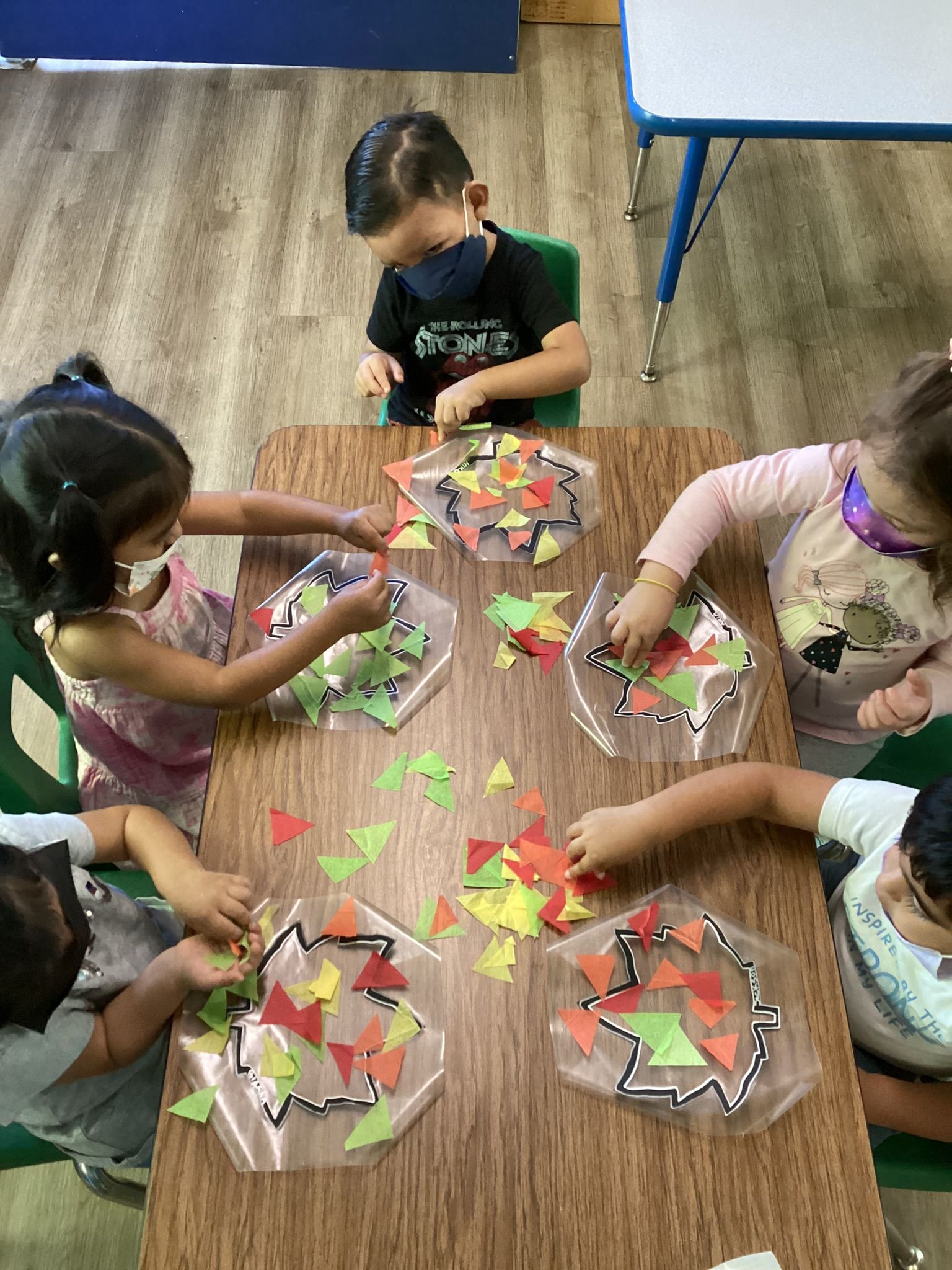 Children at a table, working on a craft with colorful triangle cutouts and clear plastic shapes.