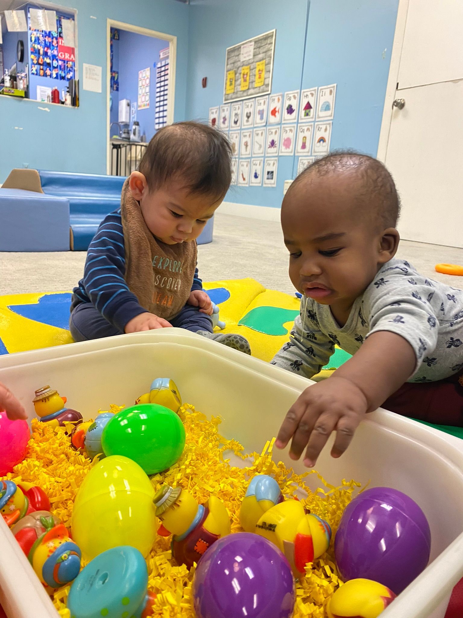 Two babies playing with Easter eggs in a classroom setting. One wears a bib.
