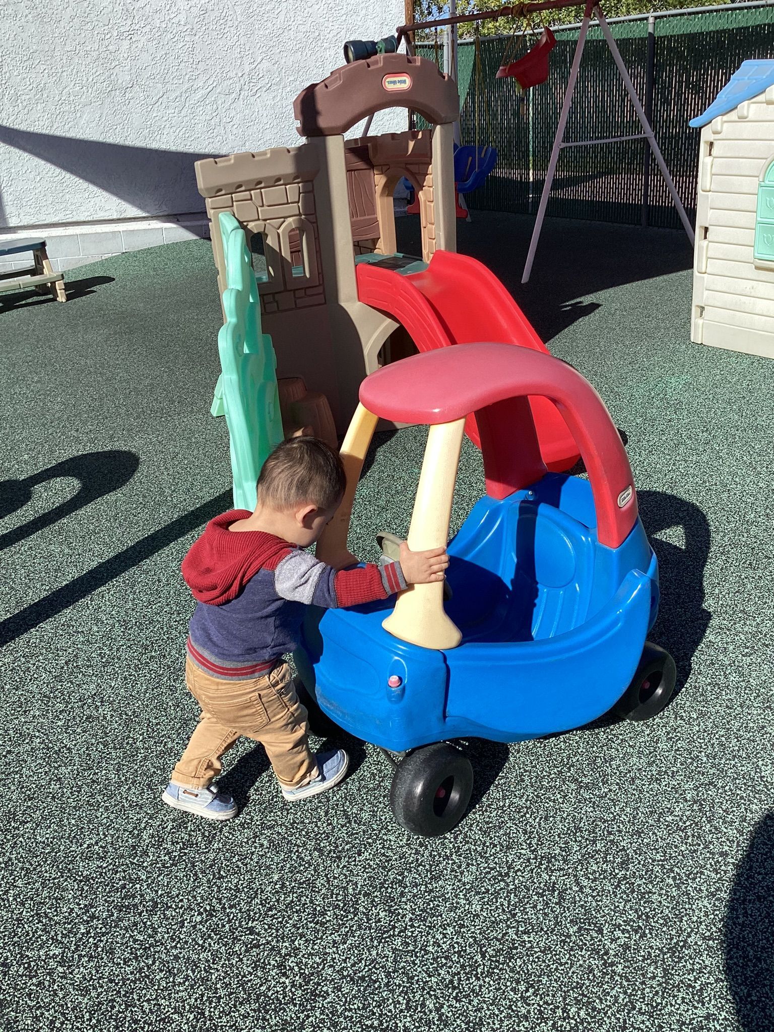 Child pushing a blue and red toy car on a playground. Brown climbing structure in the background.
