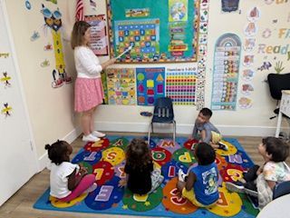 Teacher points to calendar on wall in a classroom. Children sit on rug and look toward teacher.