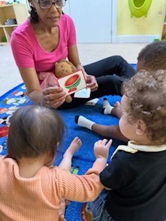 Woman reading a book with children on a blue rug, indoor setting.