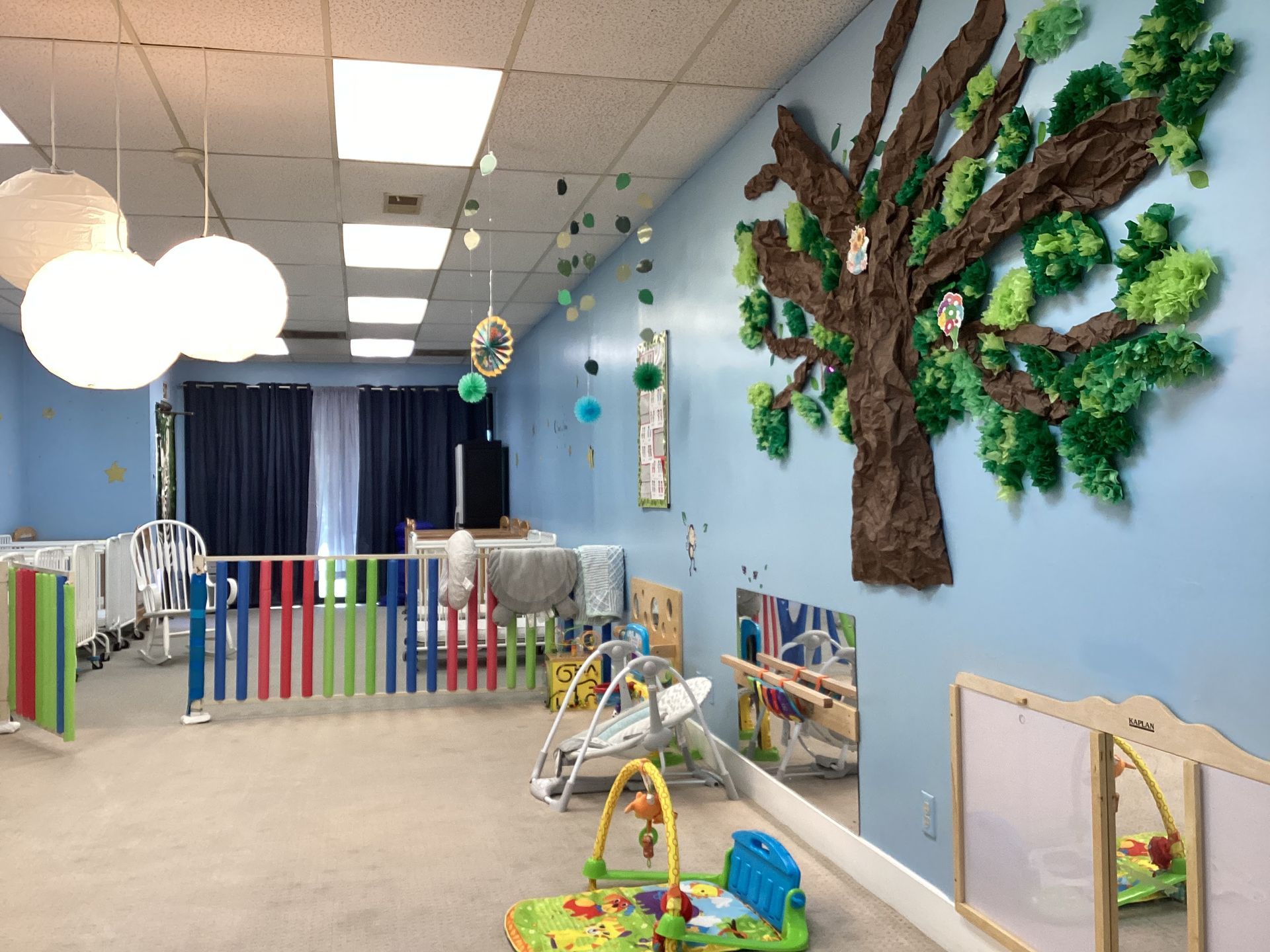 Childcare room with blue walls, decorated with a tree, paper lanterns, and a colorful playpen.