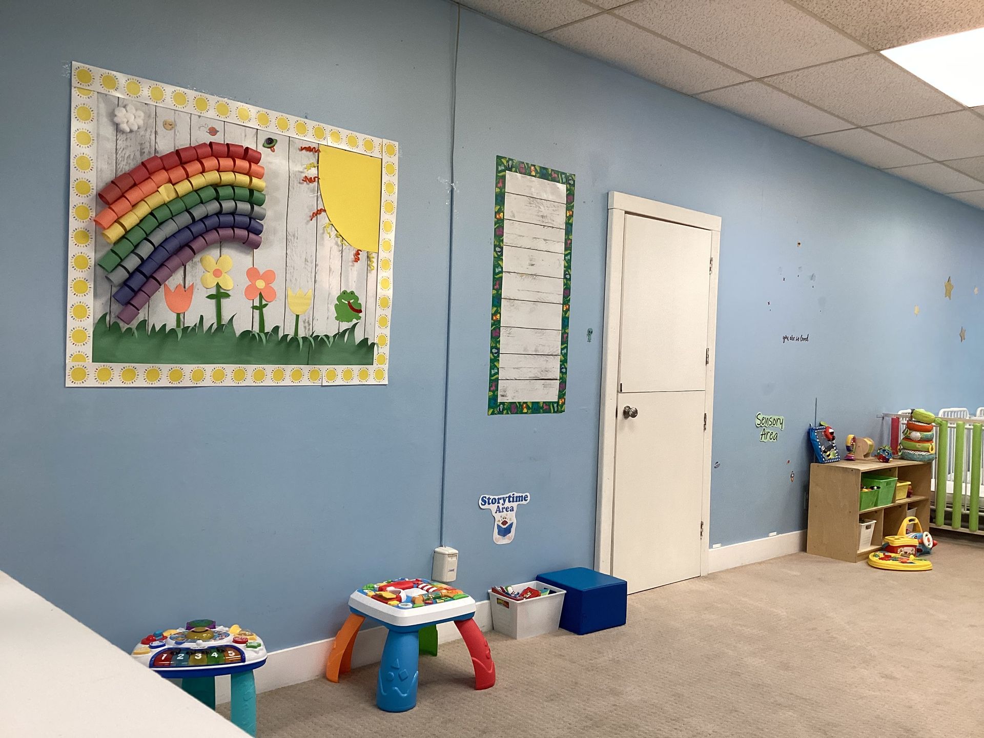 Childcare room with light blue walls, decorations, and toys. Rainbow art piece.