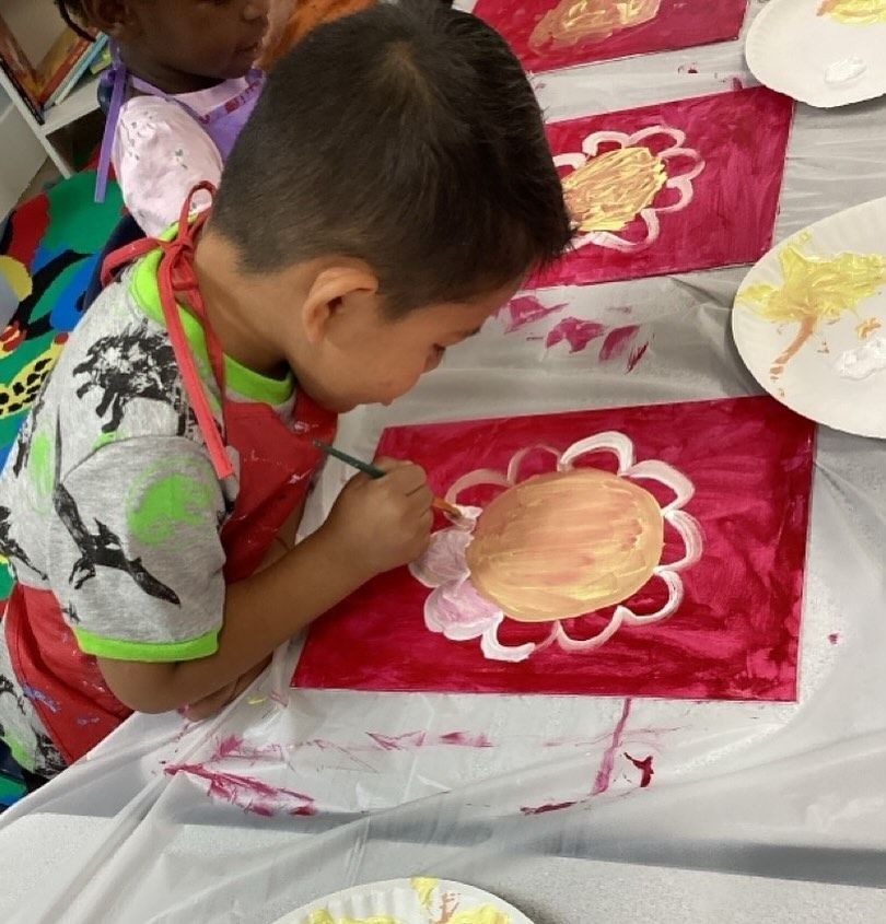 Boy painting a flower on red background, wearing apron, in a craft setting.