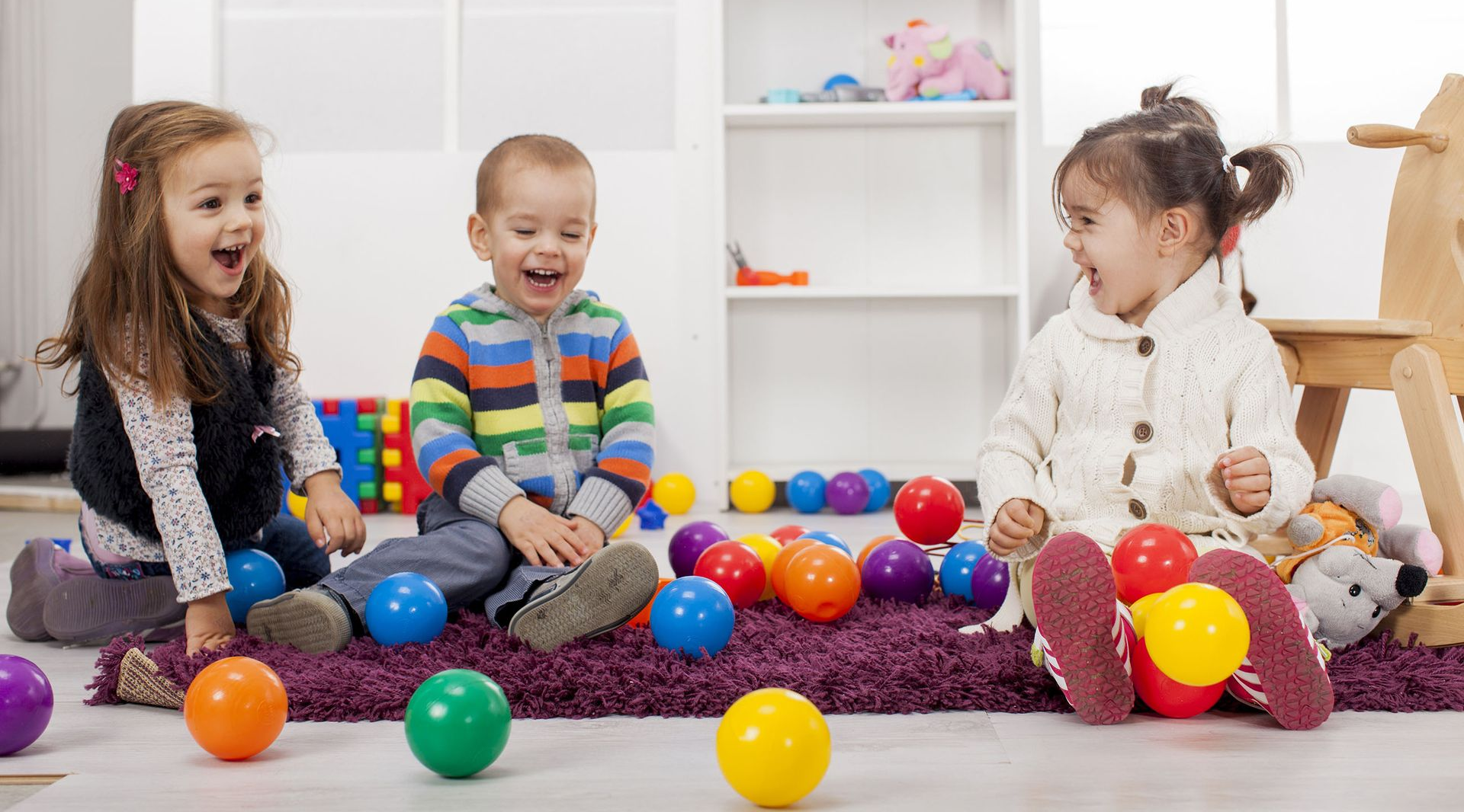 Three children laughing while playing with colorful plastic balls on a purple rug in a bright, indoor setting.