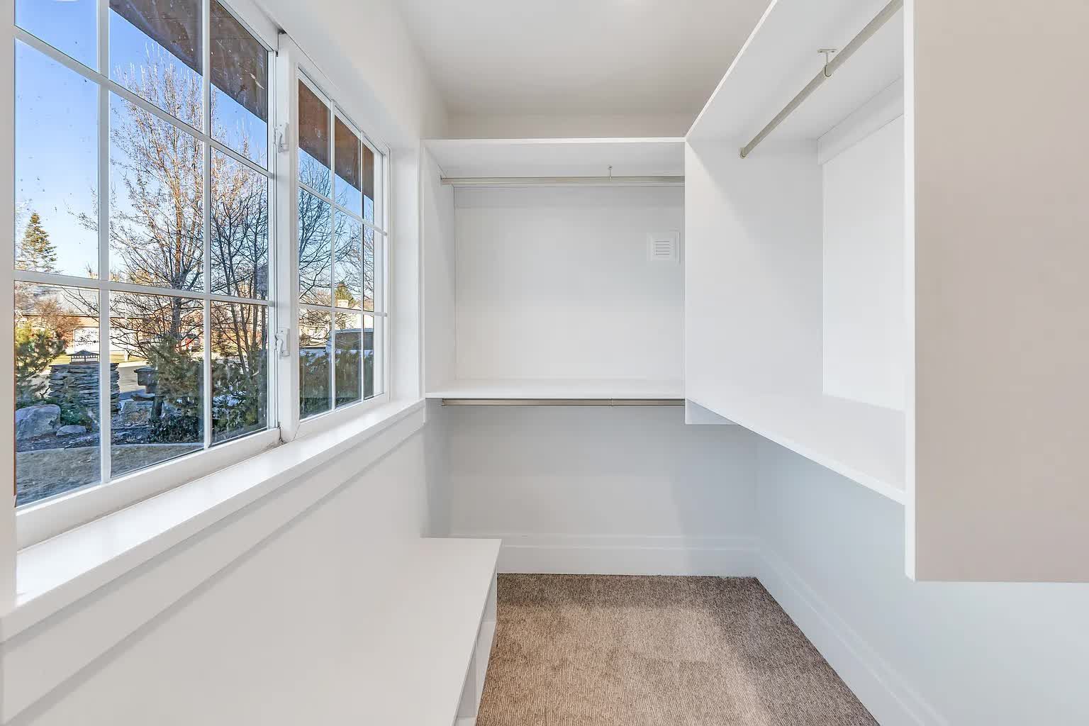 Empty white walk-in closet with shelves, a hanging rod, a window, and a bench.