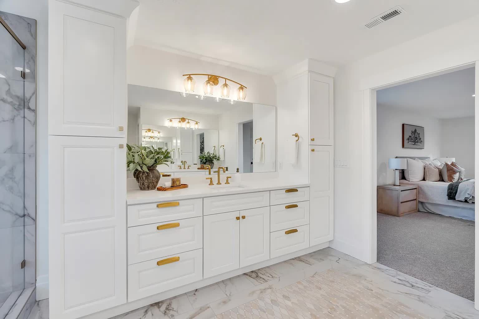 White bathroom vanity with gold hardware, mirror, and access to bedroom.